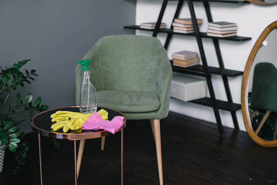 A neatly arranged living room corner featuring a light green upholstered armchair with wooden legs, positioned on a dark wooden floor. In front of the chair is a round black table with a rose gold rim