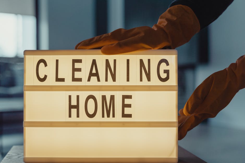 A cleaning brush with natural bristles and a wooden handle, hanging on a hook on a plain light grey wall. The brush is stored inside a light blue dustpan, which is also hanging on the hook. The wall s